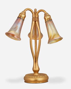 A Tiffany Studios three-light lily table lamp: Circa 1902-1919; New York, NY Each shade signed: L.C.T. / Favrile; Base signed: Tiffany Studios / New York / 319 The gilt-bronze base with a ribbed foot and tri-form stem issuing a drop cluster