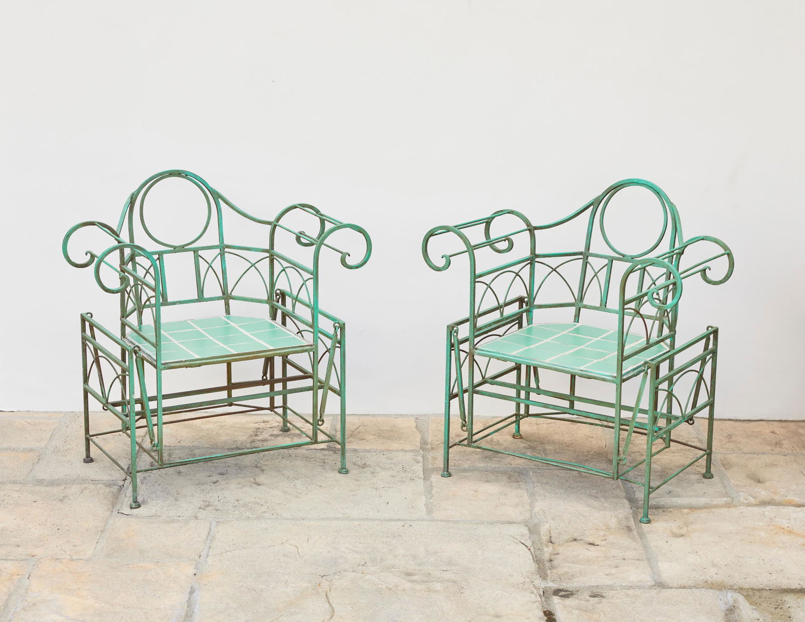 A pair of Art Deco wrought iron garden glider chairs: Circa 1930s Each verdigris patinated wrought iron chair with a pivoting gilder seat with inset green California tiles, 2 pieces Each: 34.75" H x 33.75" W x 23" D Provenance: Property from an impor
