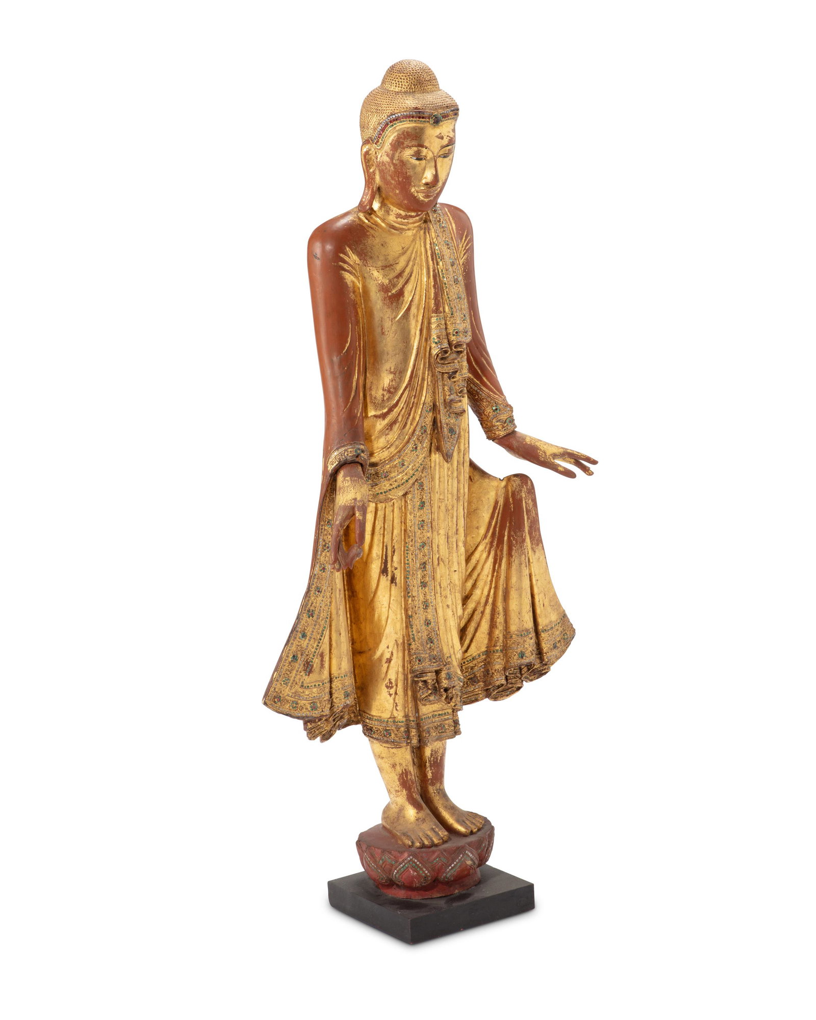 A giltwood Burmese Mandalay Buddha: 19th Century The carved giltwood standing figure depicting a youthful Buddha in elaborately folded robes trimmed with inset mirrored and glass jewels 51" H x 23.5" W x 11" D
