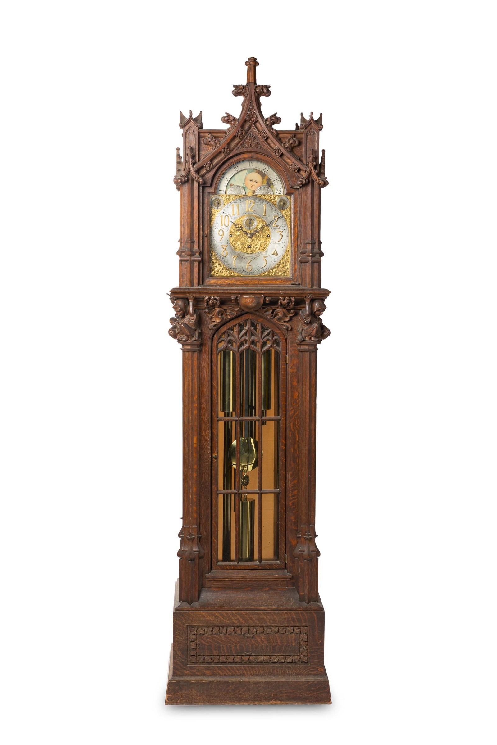 A Gothic-style tall case clock: 20th century With a silvered metal dial with brass Arabic numeral hour markers and minute track, cut iron hands, a calendar, moon face, and a decorated brass face plate, set in a Gothic-style carved w