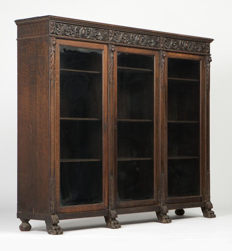 An American Renaissance Revival oak bookcase: Circa 1900, the rectangular top with molded edge over an egg-and-dart cornice above an acanthus and shield-carved frieze over three hinged glazed doors, each enclosing adjustable shelves, separated by