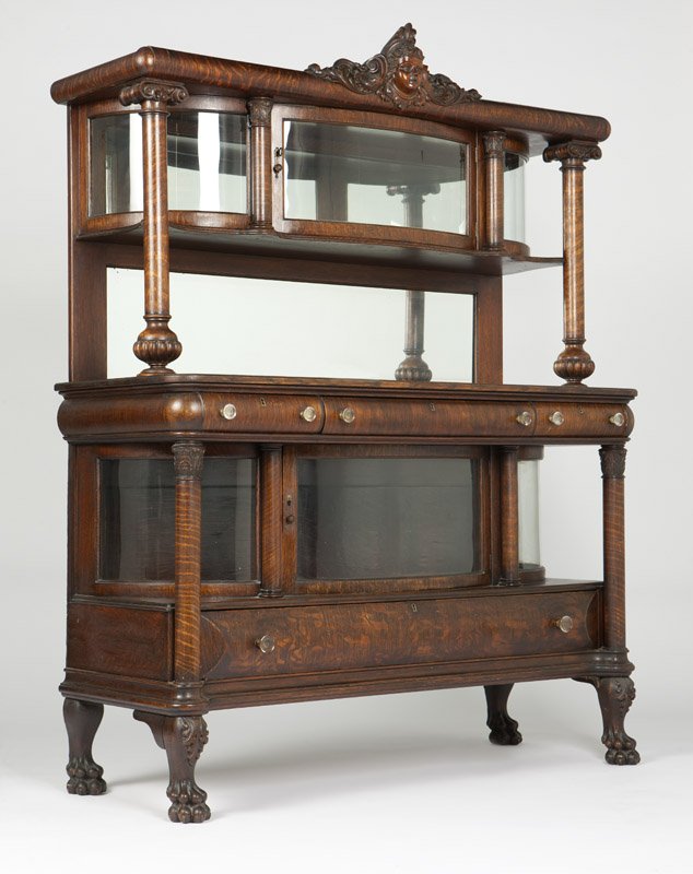 An American quarter-sawn oak buffet: Circa 1900, the rounded cornice centering a carved mask flanked by foliate scrolls over freestanding Ionic columns flanking a glazed serpentine-front mirror-backed cupboard with a central hinged door 
