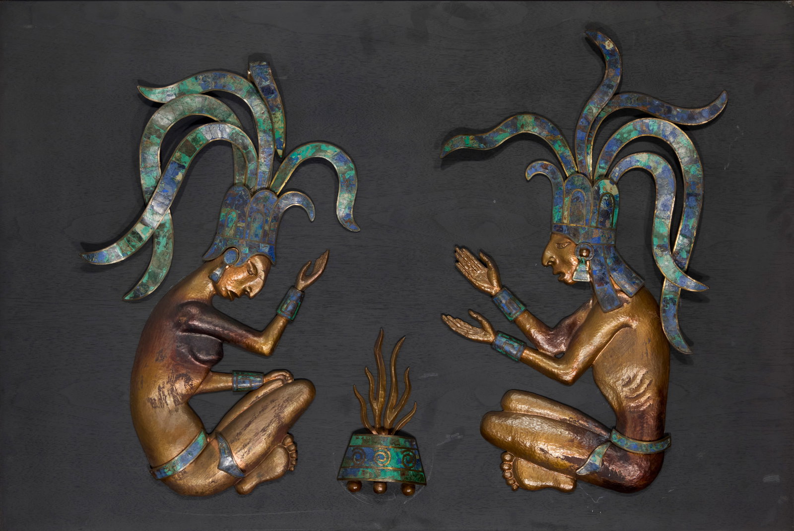 A Los Castillo Mayan/Aztec style metal and hardstone wall panel: A Los Castillo Mayan/Aztec style metal and hardstone wall panelMid/late 20th centuryUnmarkedThe hand-wrought mixed metal figures depicted seated by a fire, with inlaid azurite-malachite accents, mount