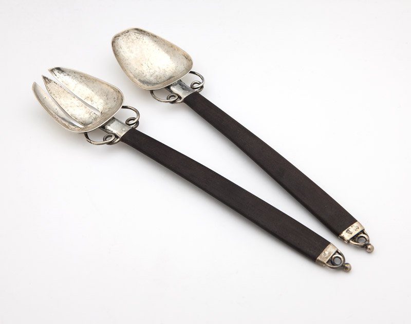 A William Spratling sterling silver salad set: Circa 1951 - 1956, each with maker's mark and Eagle mark, each with hand-hammered trapezoidal body issuing a plain ferrule and flanking wirework curlicues joined to a wooden handle terminating in a co