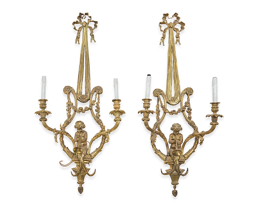 A pair of French bronze wall sconces: A pair of French bronze wall sconces 20th century Each with bow and ribbon above central trumpeter putto, garlands, and a pinecone drop finial, issuing two lights with faux candle fixtures, electrifie