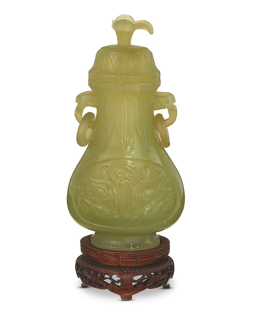 A Chinese carved jade lidded vessel 20th century Carved with Taotie mask to body, flanked by: A Chinese carved jade lidded vessel 20th century Carved with Taotie mask to body, flanked by handles with ring accents and a floriform finial, raised on a hardwood stand Jade: 6" H x 3" W x 1.875" D; 