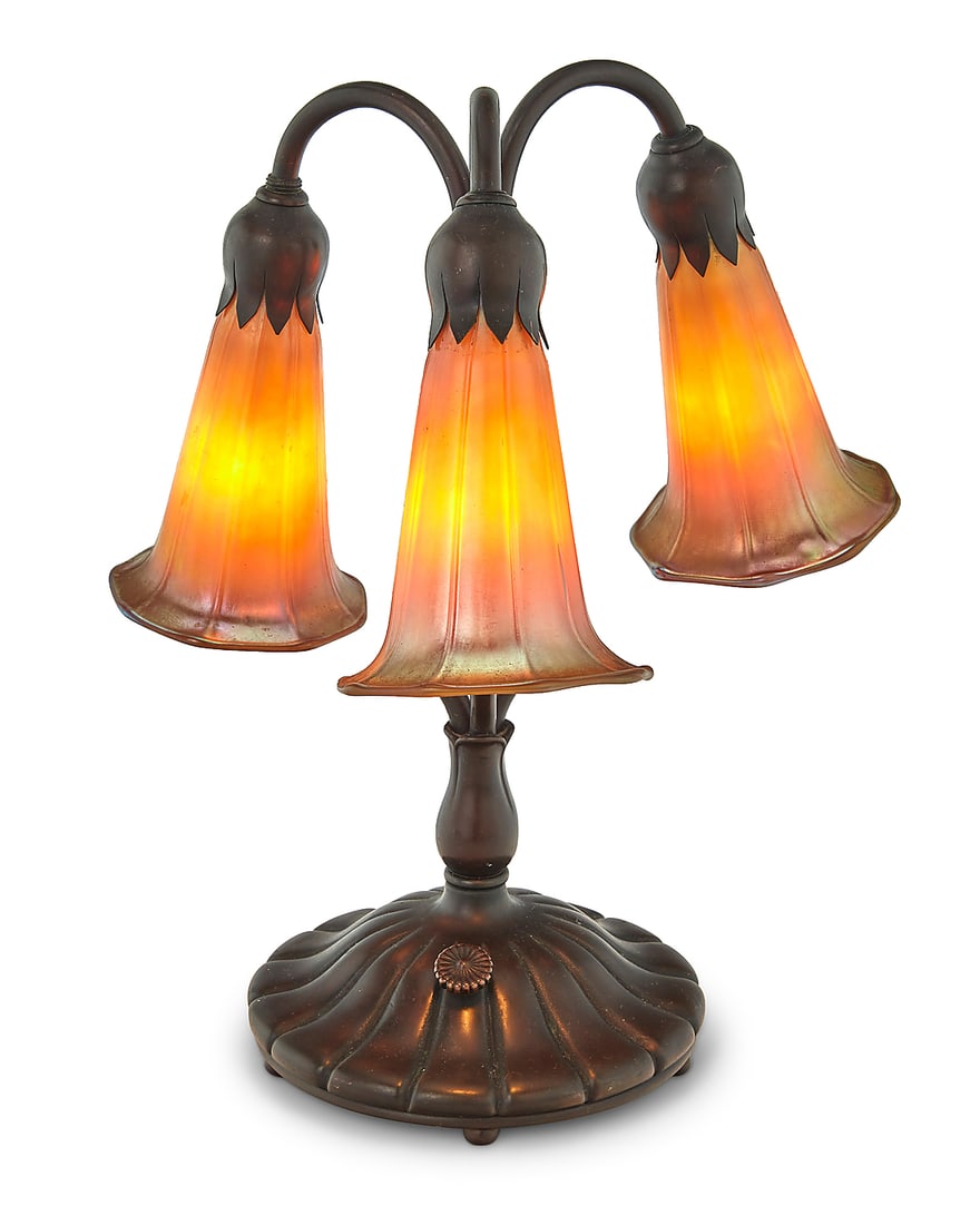 A Buffalo Metal Works Tiffany-style lily lamp: A Buffalo Metal Works Tiffany-style lily lamp 1976 The patinated copper alloy base issuing three lights, each with iridescent gold glass lily-form shade, electrified Incised with date to underside of