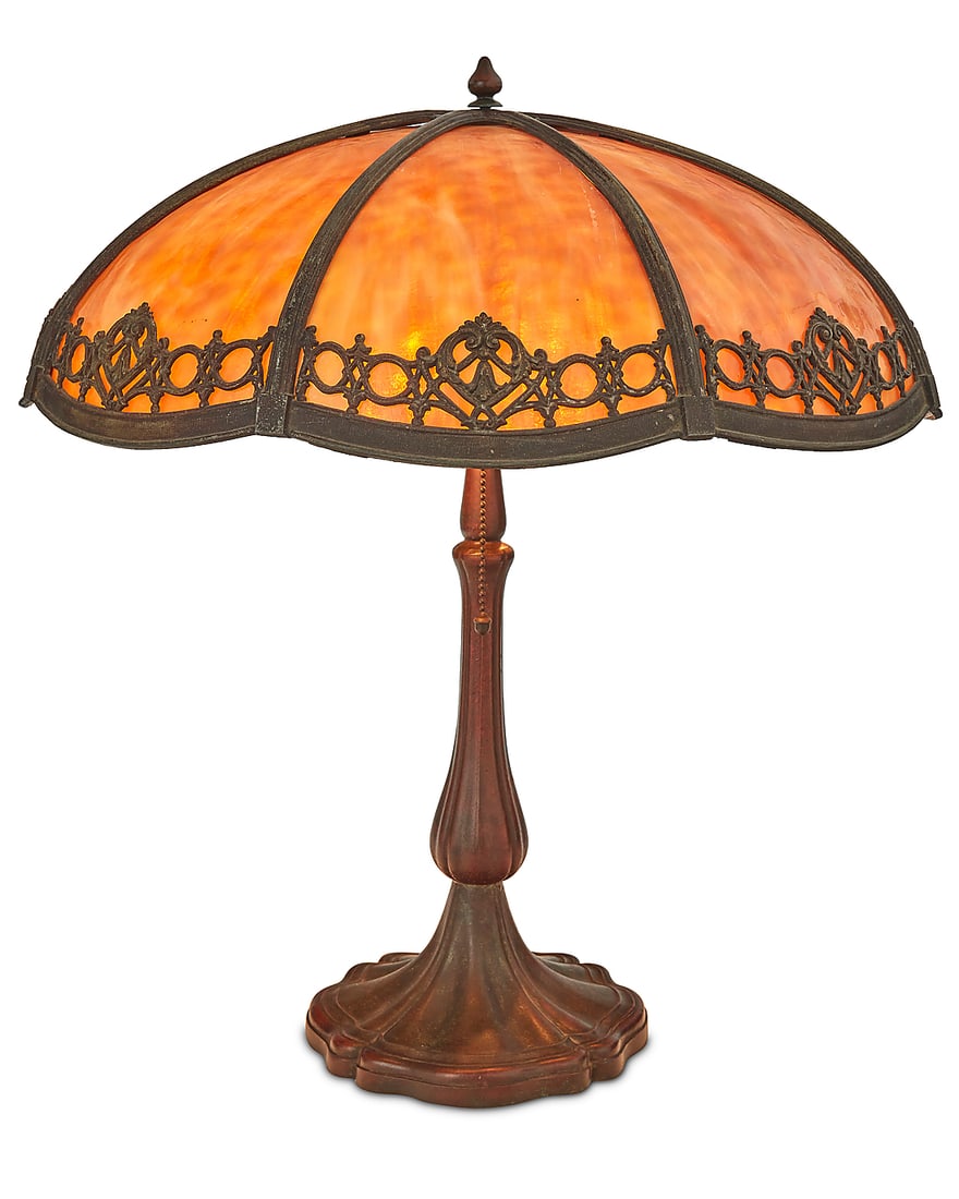 An American mottled glass and metal overlay table lamp: An American mottled glass and metal overlay table lamp Early 20th century, possibly Handel Amber slag glass shade with decorative metal overlay design, raised on a patinated metal four-lobed base
