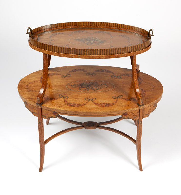 An English painted satinwood veneer tea table: Late 19th / early 20th century, the oval top tier centering a trophy within a ring of pearls surrounded by ribbon-tied floral swags, fitted with a removable glass-bottomed tray with banded sides, rais