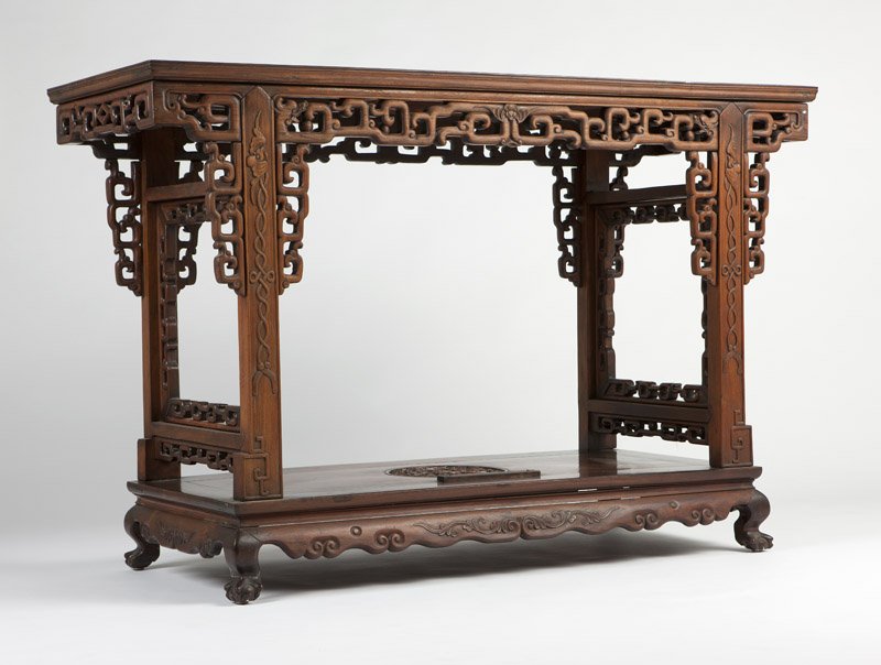 A Chinese carved wood altar table: Late 19th century, the rectangular top with inset panel over a pierced scrollwork frieze and spandrels, the frieze centering a bat and with a bird's head to each corner, raised on square supports, the