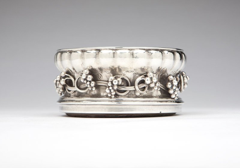 A Georg Jensen 'Grape' .830 silver wine coaster: 1919, Copenhagen, designed in 1918 by Georg Jensen, with maker's mark, date mark and .830S mark, pattern number 229, with lobed border over applied grapevine, with a grooved wooden base, 2.75'' H x 5.