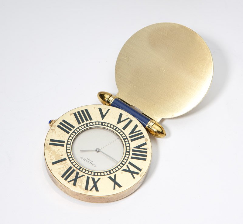 A Cartier enamel and gold-toned travel clock: Signed Cartier, Paris #357101302, the round white dial with black enamel hands, surrounded by a gold-toned case with black enameled Roman chapters and a blue stone wind stem with blue enamel stand, 14