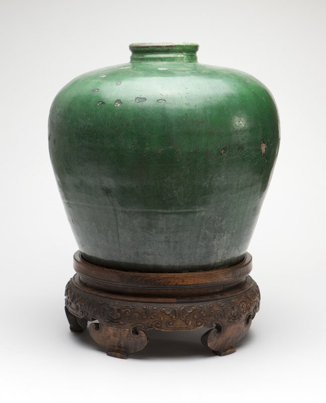 A large Chinese earthenware storage jar: 18th or 19th century, unmarked, the short circular neck over a high-shouldered body, covered with camellia leaf-green glaze, with a carved wooden base, 18'' H x 19'' Dia., est: $1000/1500