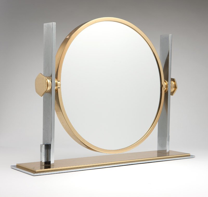 An Art Deco style chrome & brass vanity mirror: 20th century, the circular two-sided mirror plate within a conforming swivel-mount surround suspended between rectangular uprights mounted on a stepped rectangular base, 19.25'' H x 25.75'' W x 6'' D,