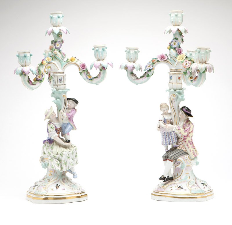 A pair of Meissen porcelain candelabra: Late 19th / early 20th century, underglaze blue crossed swords marks, each with a shaped rocaille-molded base painted with birds and butterflies, surmounted by two figures (one with a man and child, t