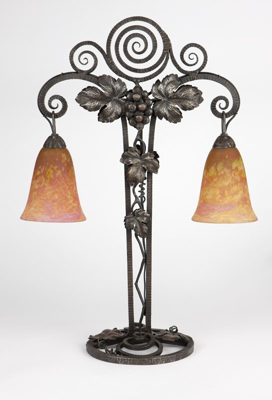 An Art Deco glass & wrought iron table lamp: The base circa 1920, cast with spirals and grapevines, suspending twin bell-shaped shades of mottled yellow and pink, each etched ''Daum / Nancy'' with cross symbol, electrified, 24.5'' H x 17.5'' W x