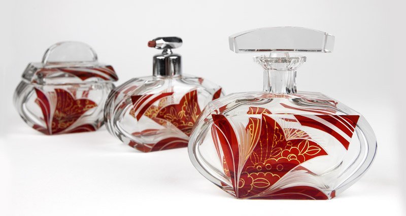 An Art Deco ruby & clear glass vanity set: Circa 1935, comprising: a covered jar (4'' H x 4.5'' W x 3'' D), a perfume bottle with stopper (4.5'' H x 5.25'' W x 2'' D), and a perfume bottle with atomizer (4.25'' H x 5.25'' W x 2'' D), each with