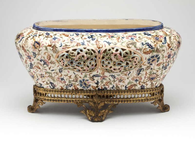 A Continental Persian-style porcelain jardiniere: Late 19th/early 20th century, the tapering high-shouldered oblong body polychrome-painted and gilt-highlighted with scrolling foliage and flowers on a white ground, each face centering a raised pierce
