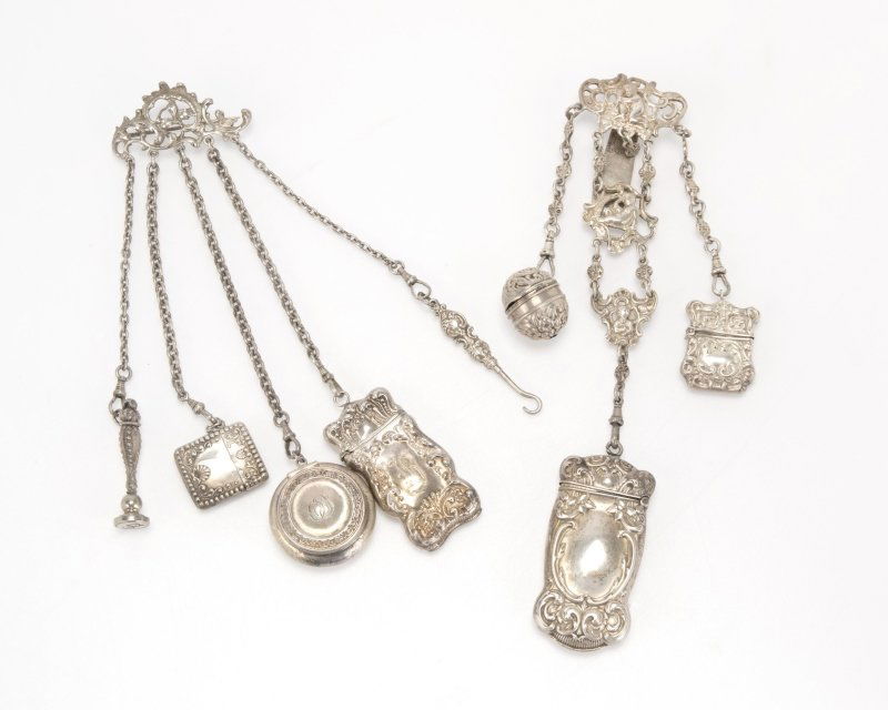 A group of two antique silver chatelaines: Circa 19th century, the first chatelaine a brooch with 5 chains suspending a seal of a lower case ''b'', a small needlecase, a coin capsule monogrammed ''OS'', a matchstrike monogrammed ''B'', and a g