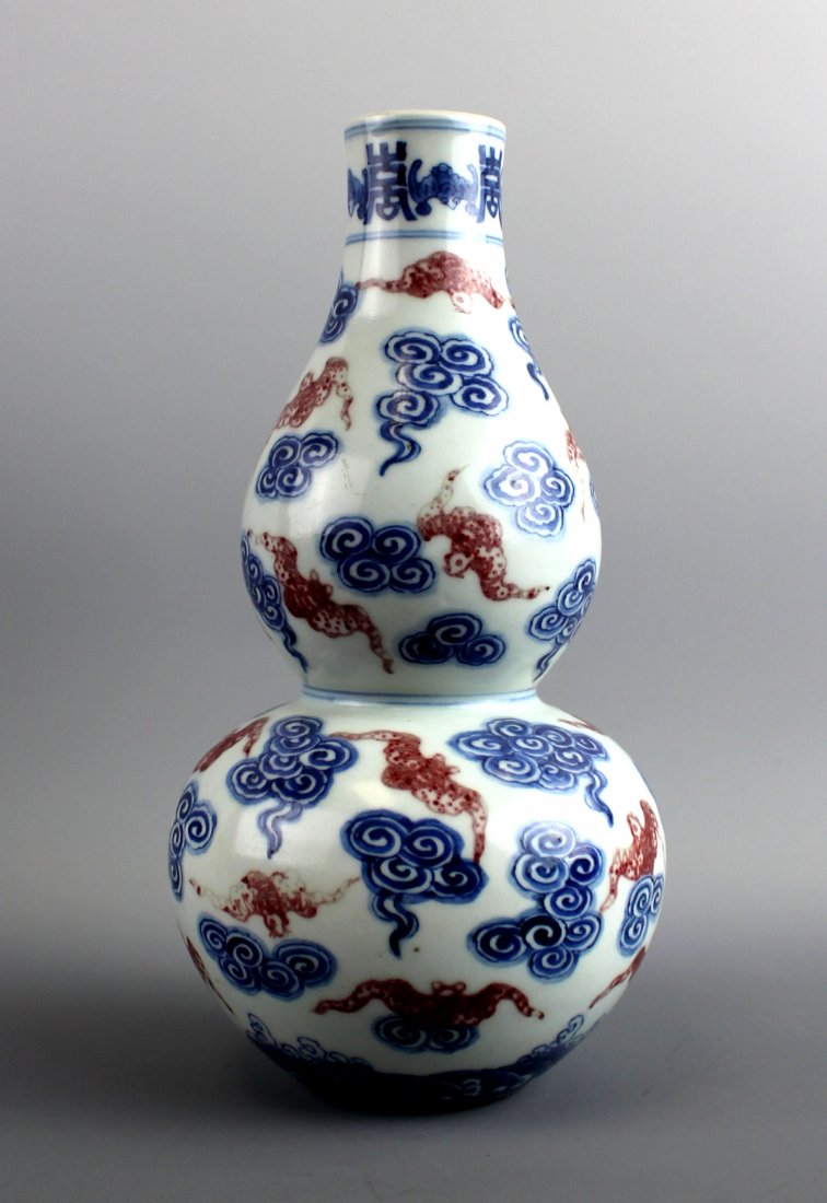 A Chinese Underglaze Copper Red and Blue Double-Gourd: The elegant double-gourd form vase features red bats painted in underglaze copper red flying among blue clouds. A band of bats alternating with shou (Longevity) characters borders the top and another 