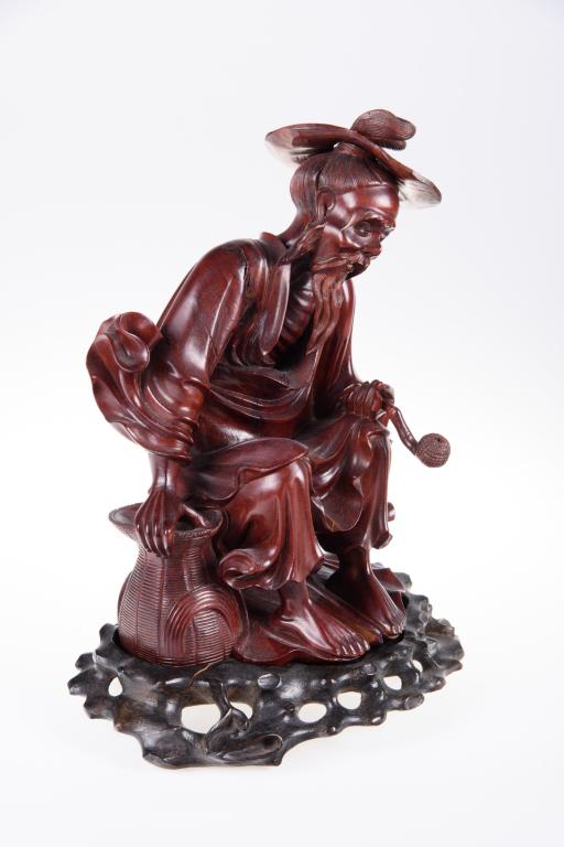 Carved Wood Asian Figure: Man seated, wearing straw hat, holding a pipe (chip to stem) and with one hand resting on a basket. Age checks to wood. On a fitted wood base. Measures approximately 11" in height with base.