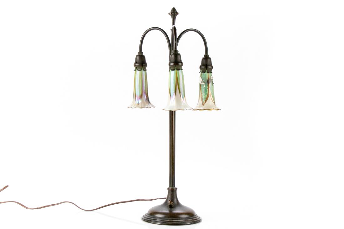 Quezal Glass And Bronze Arts And Crafts Lamp: A three arm bronze lamp with a tiered circular base, rod standard, and flower bud finial. The three arched arms hold the three marked pendant Quezal glass flower form shades. They are in a green and w