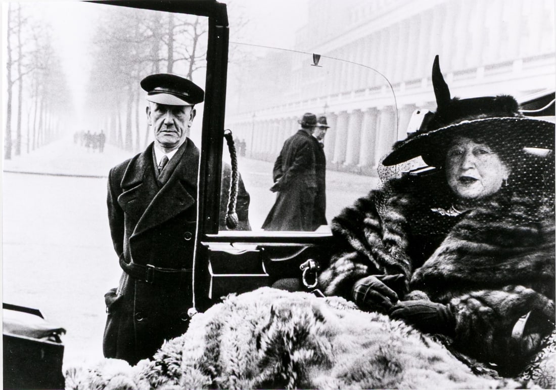 Inge Morath (Austrian, 1923-2002) "Mrs. Eveleigh Nash..." Signed Gelatin Silver Print: "Mrs. Eveleigh Nash, Buckingham Palace Mall, London, 1953" gelatin silver print depicting a woman in ornate fur and a feathered hat sitting in a car while men walk by urban architecture and a tree-lin