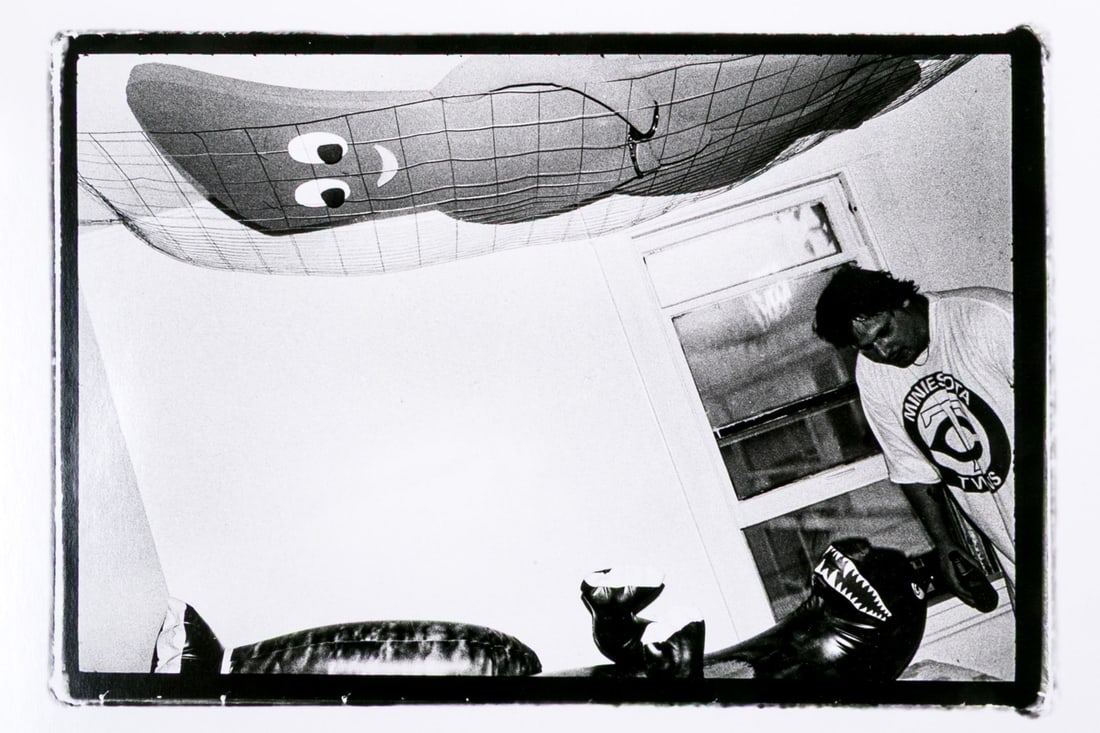 Ramon J. Muxter (American, 1945-2007) "Robert, 1983" Signed Gelatin Silver Print: Ramon J. Muxter (American, 1945-2007) "Robert, 1983" Gelatin silver print depicting a man in a room interior with a large handcuffed foam figure in a net across the ceiling and an inflatable dinosaur 