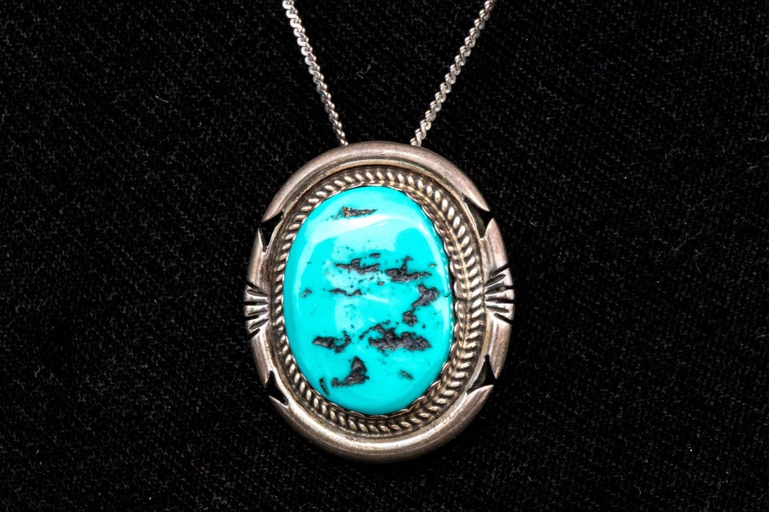 NOTABLE NAVAJO JON MCCRAY TURQUOISE PENDANT/BROOCH, WITH IHMSS HALLMARK: This beautiful Navajo sterling silver brooch was made by Jon McCray, and features a fine oval turquoise center, decorated with a rope edge and cut border frame. Accompanied by an "S" style chain, with