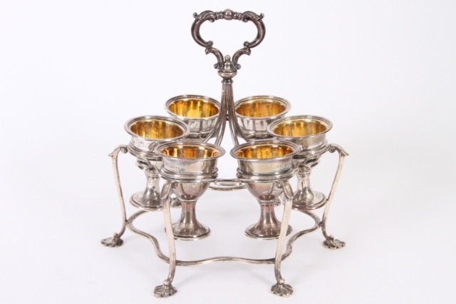 VICTORIAN STERLING EGG CUP HOLDER Ca 1857: Having six cups held in a footed stand with ornate handle, cups have gold-washed interior. Stand measures 8" diameter x 8" high, cups measure 2 1/2" high x 2" diameter. 24 T. O.