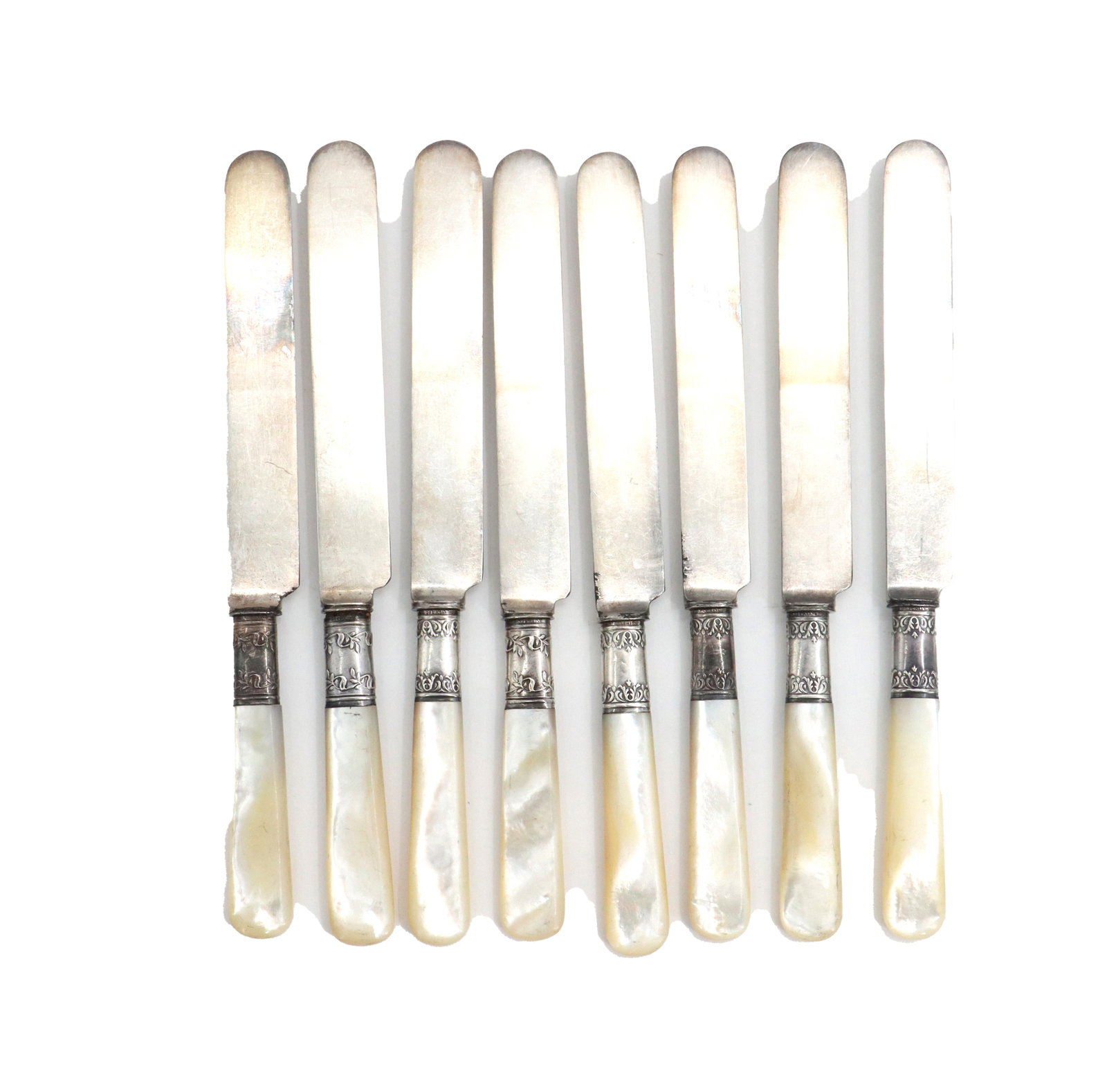 (8) Mother of Pearl Handle Butter Knives: (8) Mother of Pearl Handle Butter Knives. Mother of pearl handles are joined to the metal blades by ornate, engraved ferrules. Blades are short and blunt tipped, typical for this style of early 