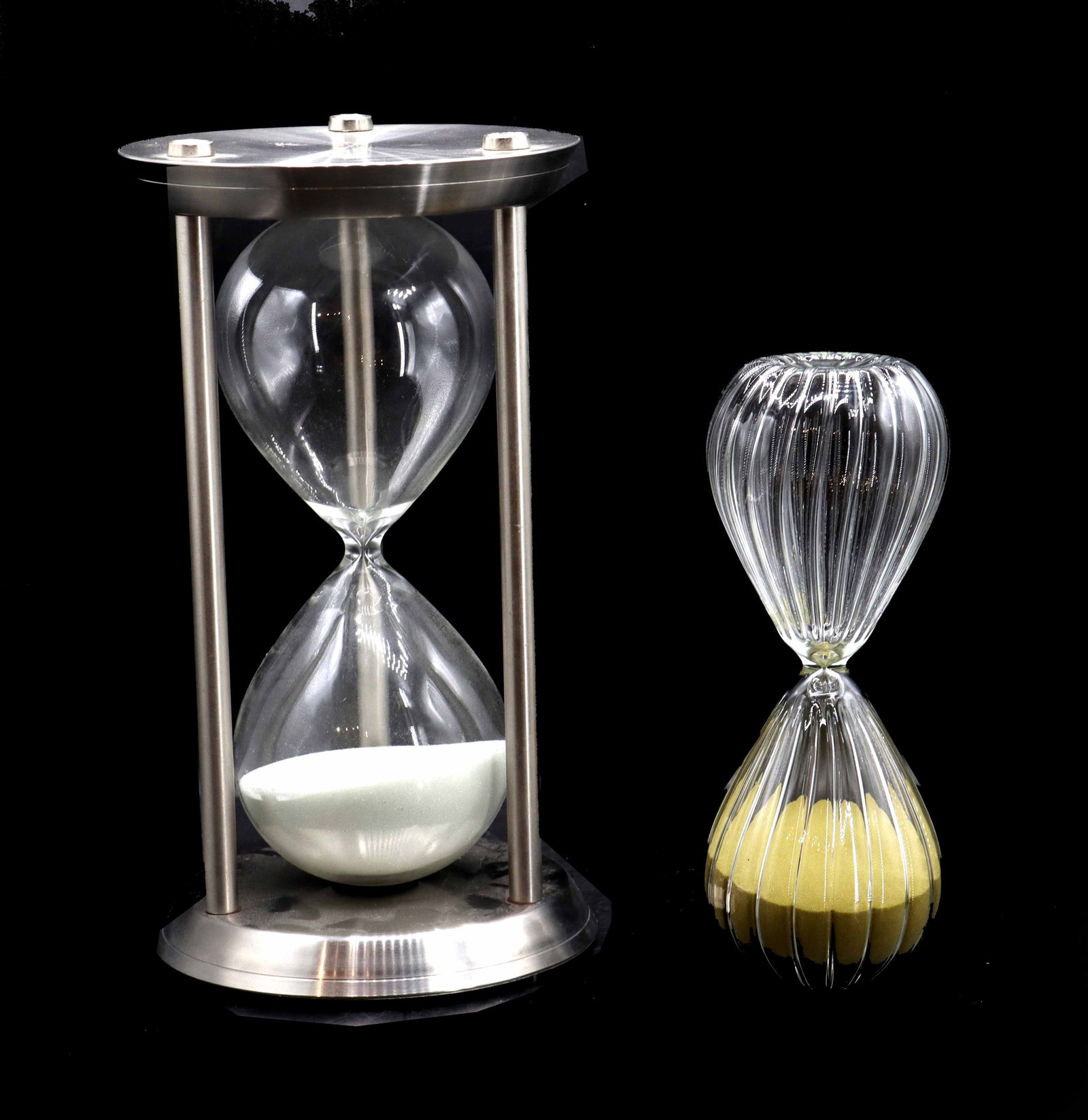 (2) Large Hour Long Hourglasses: (2) Large Hour Long Hourglasses. The bigger one is filled with white sand, and is bookended by metal bases connected by three rods. The smaller one is filled with yellow sand, and has a