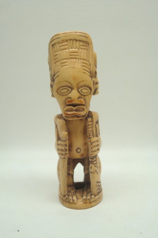Antique Carved African Ivory: Antique carved African Ivory figure depicted wearing headdress. Figure has almond shaped eye, and is in squatting position. Holding two staffs in either hand. Headdress having two small forms on both 