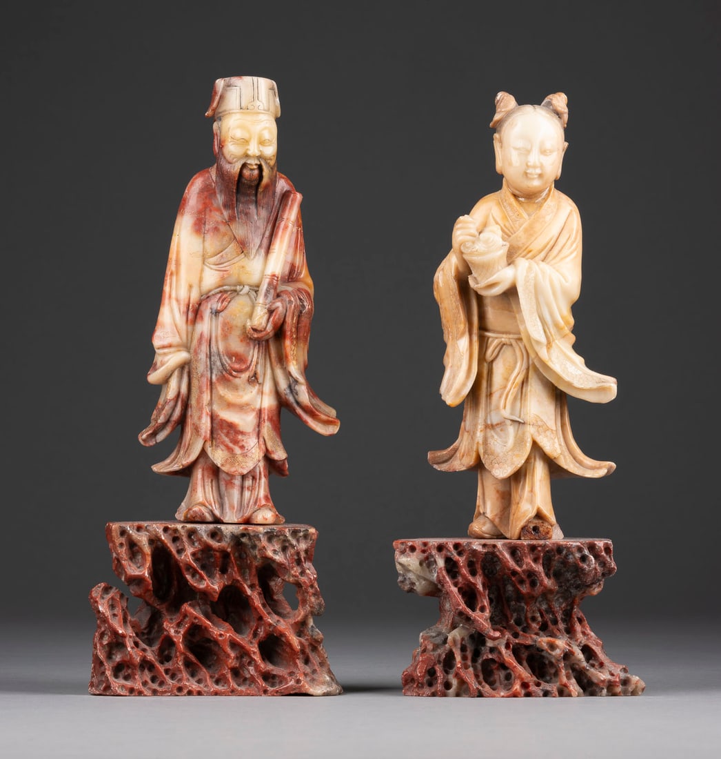 TWO SOAPTONE CARVED STANDING FIGURES OF IMMORTALS: TWO SOAPTONE CARVED STANDING FIGURES OF IMMORTALS China, Qing Dyansty Soapstone. H. 20-20.2 cm (27.3-28 cm with base). Including a figure of Zhang Guolao and a figure of He Xiangu, both standing on a