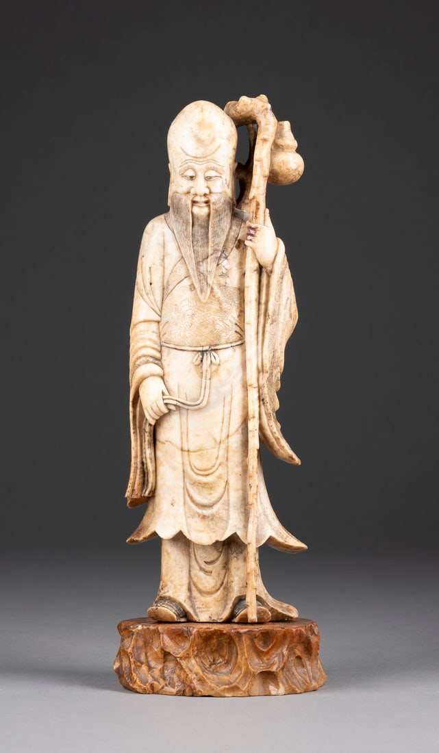 A SOAPTONE CARVED STANDING FIGURE OF SHOULAO: A SOAPTONE CARVED STANDING FIGURE OF SHOULAO China, late Qing Dyansty Soapstone. H. 28 cm (31.5 cm with base). The god of longevity is standing on a separate base in form of rock, holding a gnarled st