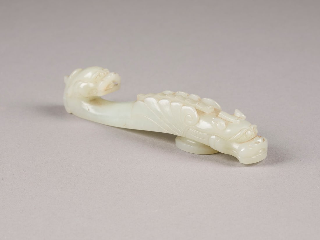 EIN GUERTELHAKEN AUS PALEZELADON-JADE: A PALE CELADON JADE BELT HOOK China, Qing Dynasty Jade. L. 11.5 cm. The terminal is carved in high relief with a chi-dragon head, the curving body further carved with another chi-dragon. With traces o