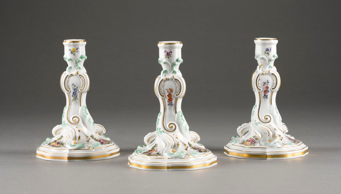 | DREI KERZENHALTER 'WATTEAU- UND BLUMENMALEREI': DREI KERZENHALTER 'WATTEAU- UND BLUMENMALEREI' Deutsch, Meissen, 2 Hälfte 20. Jh. Porzellan, polychrome Malerei, Goldstaffage. H. 18 cm. Unterglasurblaue Schwertermarke, Press- und Malernummer, Jahre