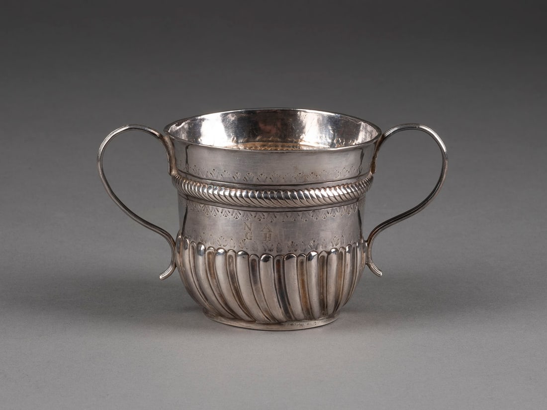 | KLEINER HENKELBECHER: KLEINER HENKELBECHER England, London, 1711 Silber, getrieben. H. 7,4 cm, 106 g. Punziert mit Garantiemarken, Meistermarke 'B' und Jahresbuchstaben 'Q'. Wandung mit Godronenfries und graviertem