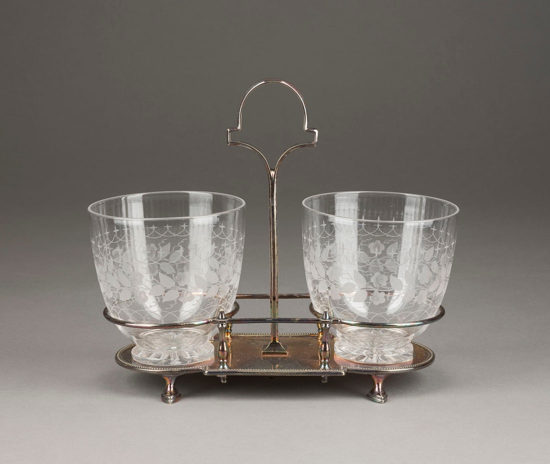 | MENAGE MIT ZWEI GLÄSERN: MENAGE MIT ZWEI GLÄSERN 20. Jh. Weißmetall, versilbert, farbloses, teils satiniertes Glas. H. 20,5 L. 24 cm. Unterseitig mehrfach punziert sowie mit Modell-Nr. '3087'. Min. Gebrauchsspuren.MENAGE