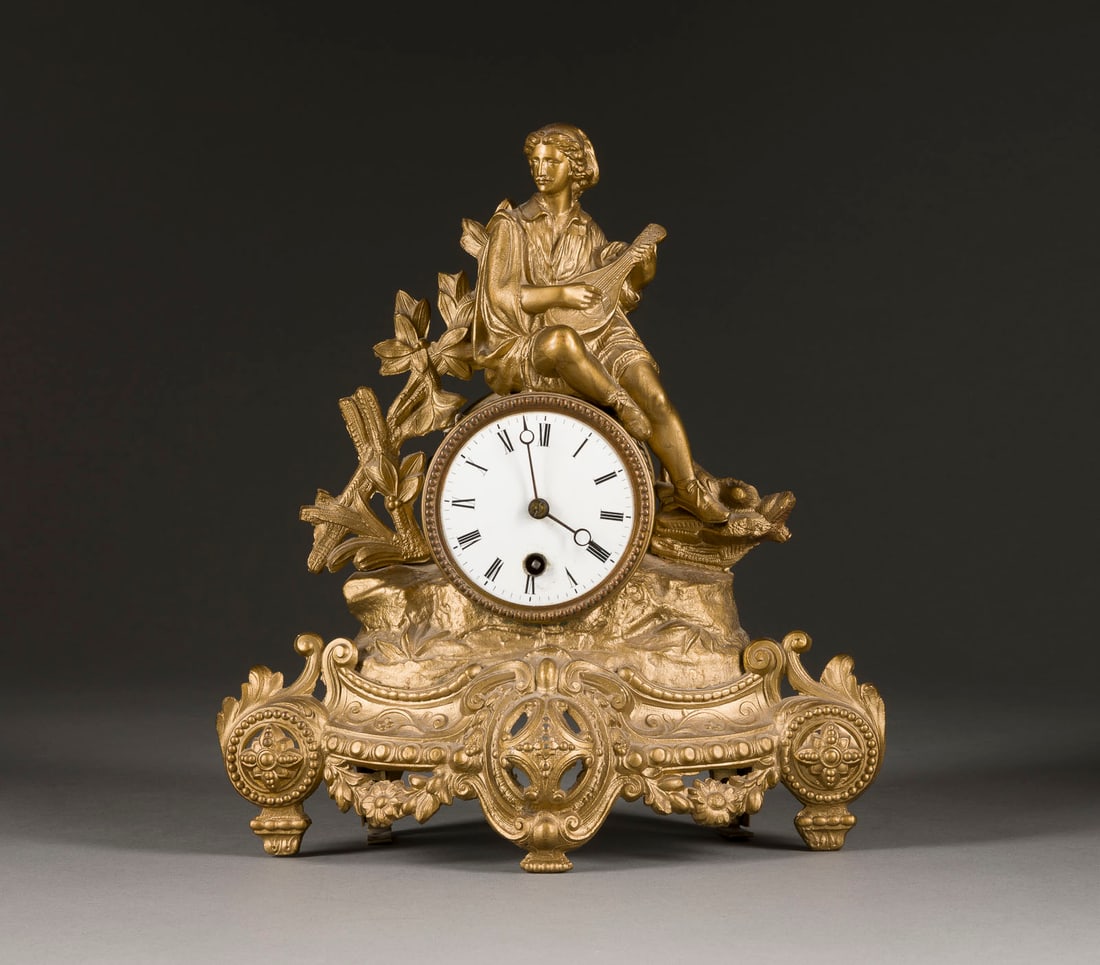 | TISCHUHR MIT LAUTESPIELER: TISCHUHR MIT LAUTESPIELER um 1900 Bronzierter Metall- oder Zinkguss, Email, Stahl. 30 x 27 x 11 cm. Über reich verziertem Sockel mit Floraldekor plastische Figur eines Lautespielers. Weißes, emailli