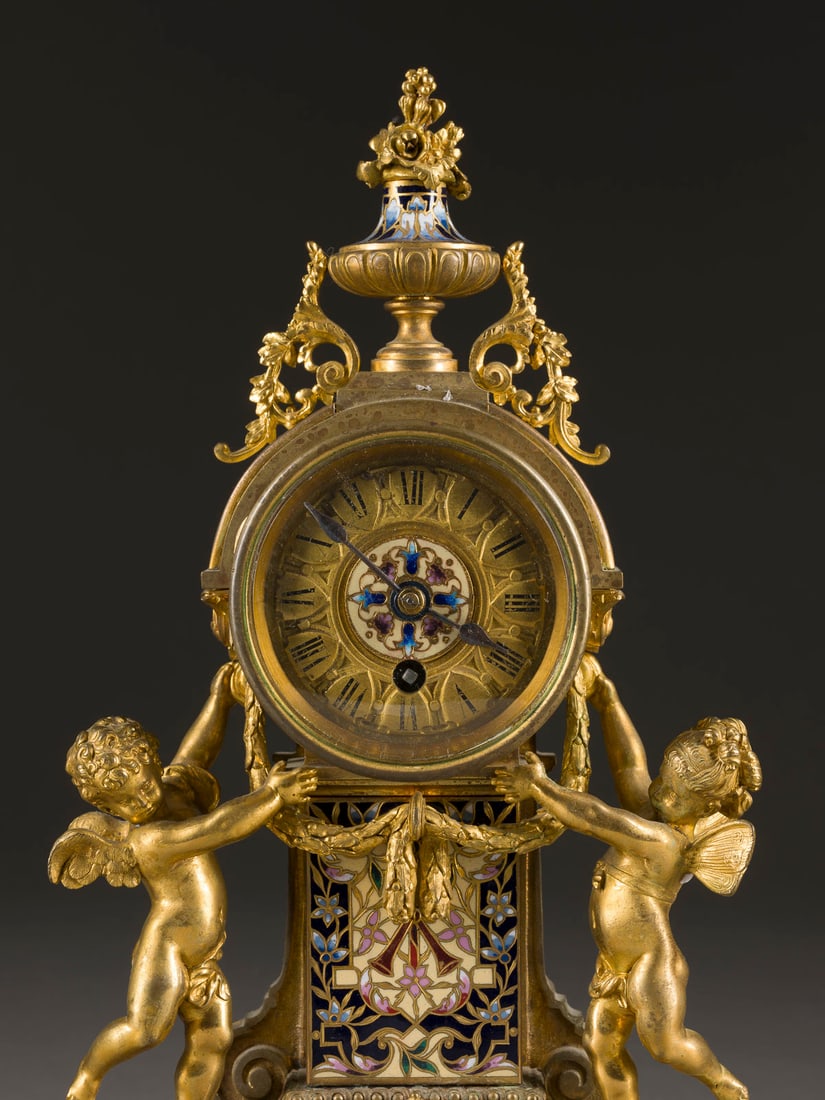 | HISTORISMUS-TISCHUHR: HISTORISMUS-TISCHUHR um 1880 Bronze, vergoldet, Email, Glas, Metall, Stahl. 28 x 14,5 x 8,5 cm. Mit polychromem Cloisonne-Email reich verzierter Korpus. Von zwei Putten flankiertes Uhrwerk und