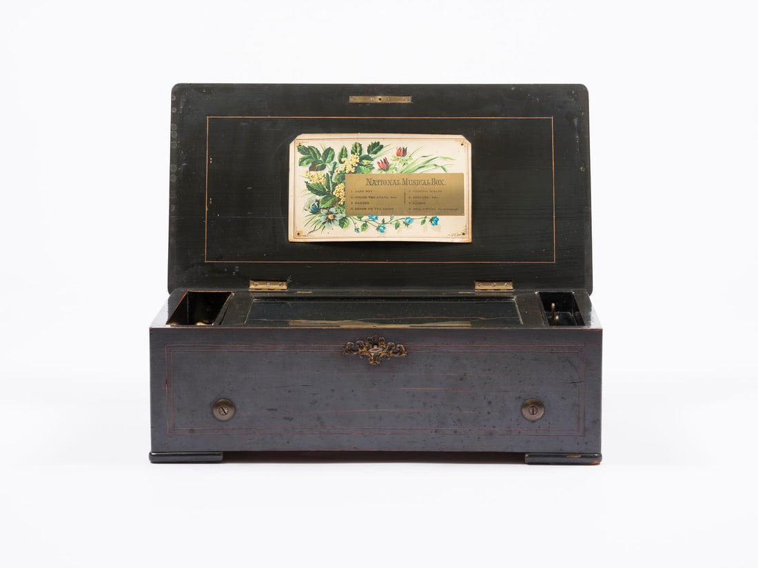 | WALZENSPIELDOSE 'NATIONAL MUSICAL BOX': WALZENSPIELDOSE 'NATIONAL MUSICAL BOX' England, um 1900 Nussbaumkasten, intarsierte, florale Ornamente, Walzenspielwerk, Metall, Messing, Glasdeckel. H. 15, B. 45, T. 24 cm. Innen Löwenmarke. Walzens