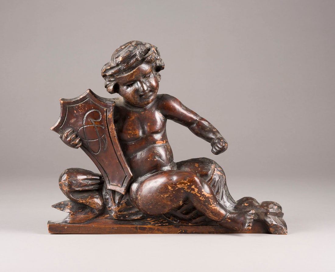 | SITZENDER PUTTO MIT WAPPEN: SITZENDER PUTTO MIT WAPPEN Deutsch, 18. Jh. Holz, plastisch geschnitzt, dunkel gebeizt. H. 33, B. 40, T. 10 cm, Gew. ca. 1,94kg. Part. best. und besch.SITZENDER PUTTO MIT WAPPEN