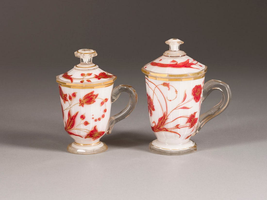 | PAAR DECKELTASSEN MIT FLORALEM DEKOR: A PAIR OF LIDDED GLASS CUPS WITH FLORAL DECOR Russian, probably Imperial Glass Factory, mid 19th century Colourless, facetted bodies, milk glass overlay, red and gold painting. Smaller cup rest.