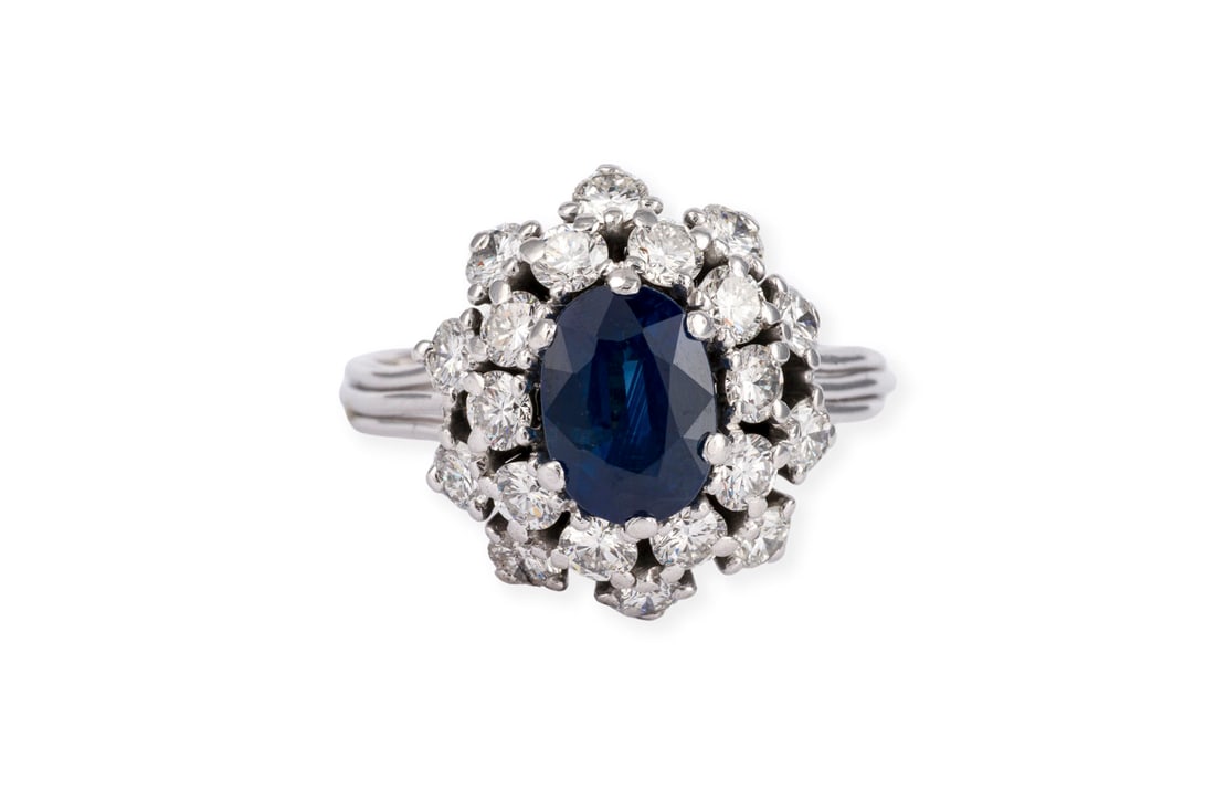 SAPHIR COCKTAIL RING: SAPHIR COCKTAIL RING WG 750 (getestet). Ringmass: ca. 58. Ringkopfmasse: ca. 16,6 x 15,1 mm. Ges.-Gew.: ca. 7,2 g. Ring, besetzt mit einem zentralen Saphir von ca. 1.85 ct. und einem doppelten Kranz