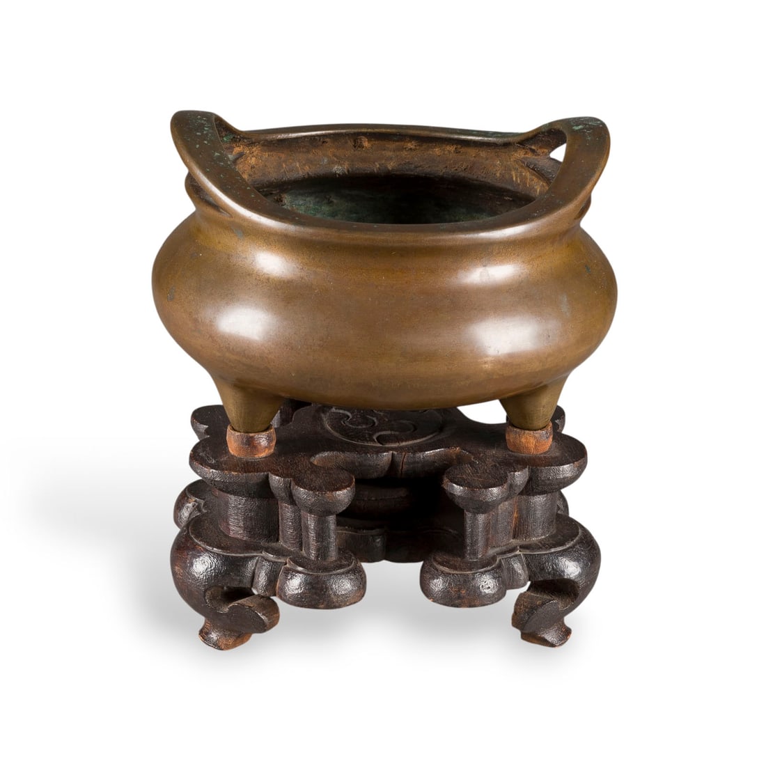EIN WEIHRAUCHBRENNER AUF DREI FUESSEN AUS BRONZE MIT HOLZSTAENDER: A BRONZE TRIPOD CENSER AND WOODEN STAND China, Qing Dynasty Bronze. H. 12.2 cm (21.5 cm with stand). The underside decorated with two chasing dragons enclosing a Xuande mark. With traces of use.EIN WE