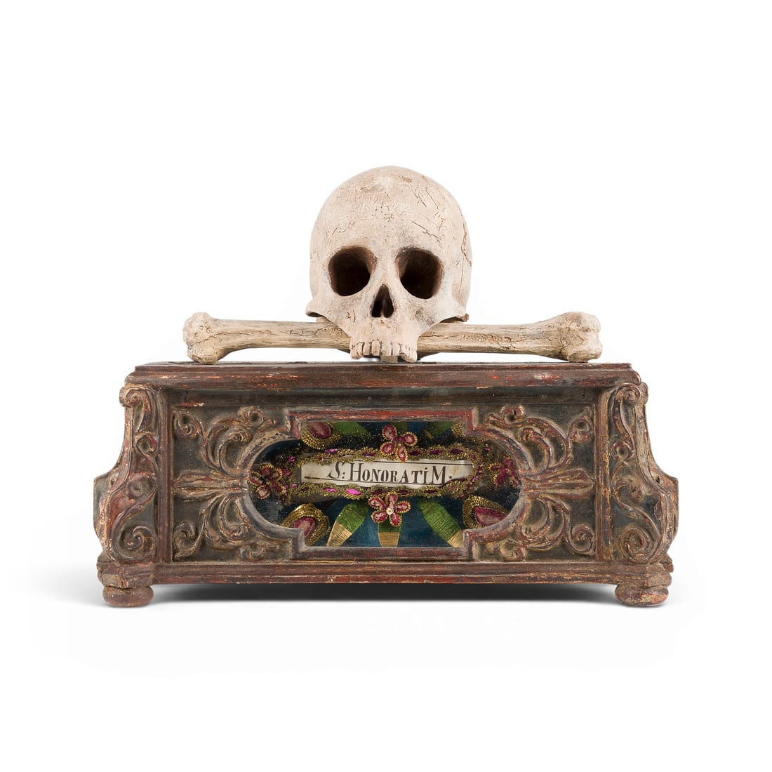 | MEMENTO MORI HAUSRELIQUIAR 'HEILIGER HONORATUS' MIT TOTENSCHAeDEL UND KNOCHEN: MEMENTO MORI HOUSE RELIQUARY 'SAINT HONORATUS' WITH SKULL AND BONES Germany or Austria, 19th century Wood, carved in high relief, polychrome finish, with display case. H. 34, W. 43, D. 16 cm; weight a