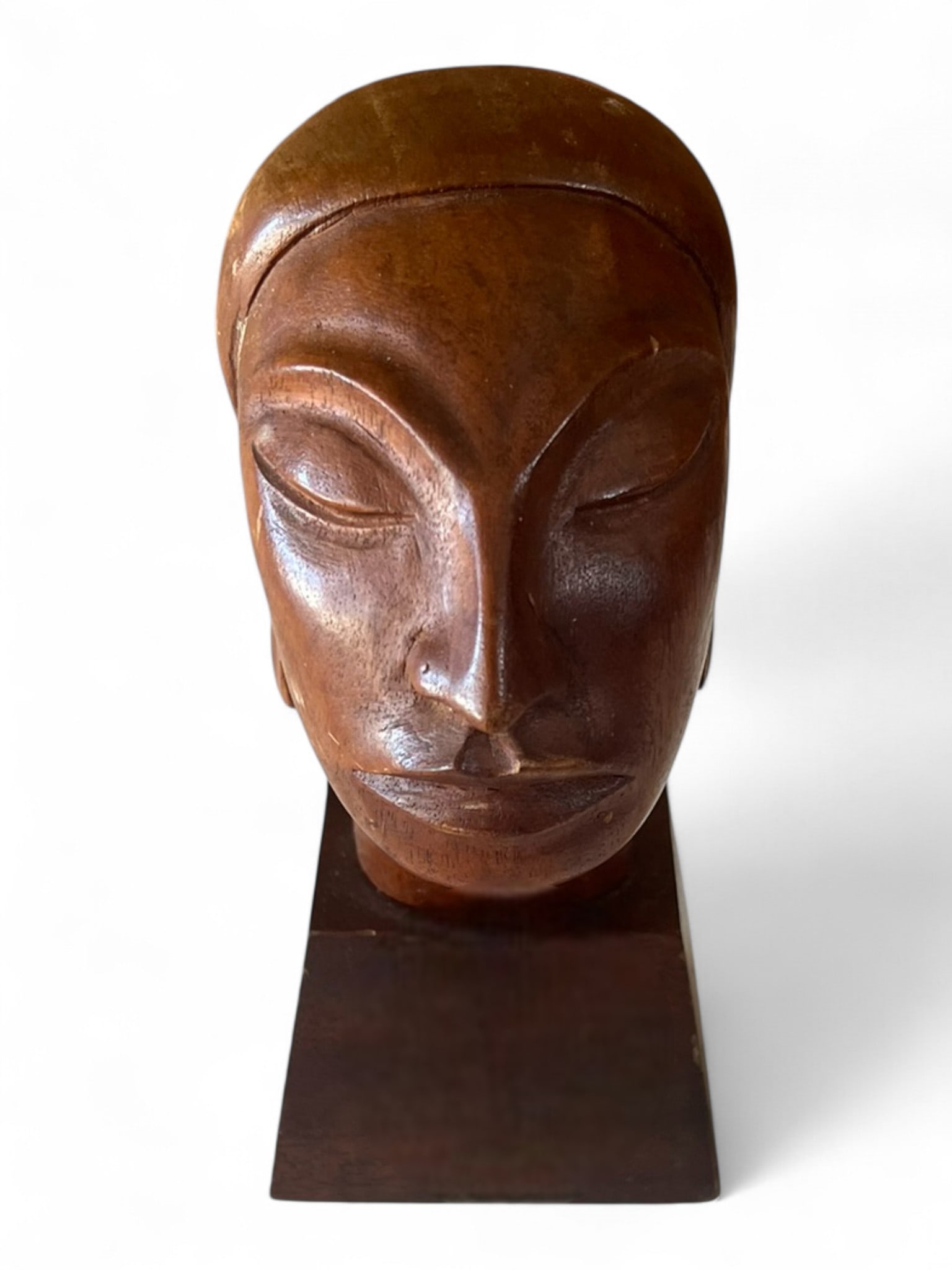 Signed J. Pinal Hand-Carved Wooden Female Bust, Latin America, mid-20th century: A finely carved and polished mahogany bust depicting a stylized female head with serene expression and smooth facial planes, the hair drawn back into a bun and mounted on a square plinth. The sculptur