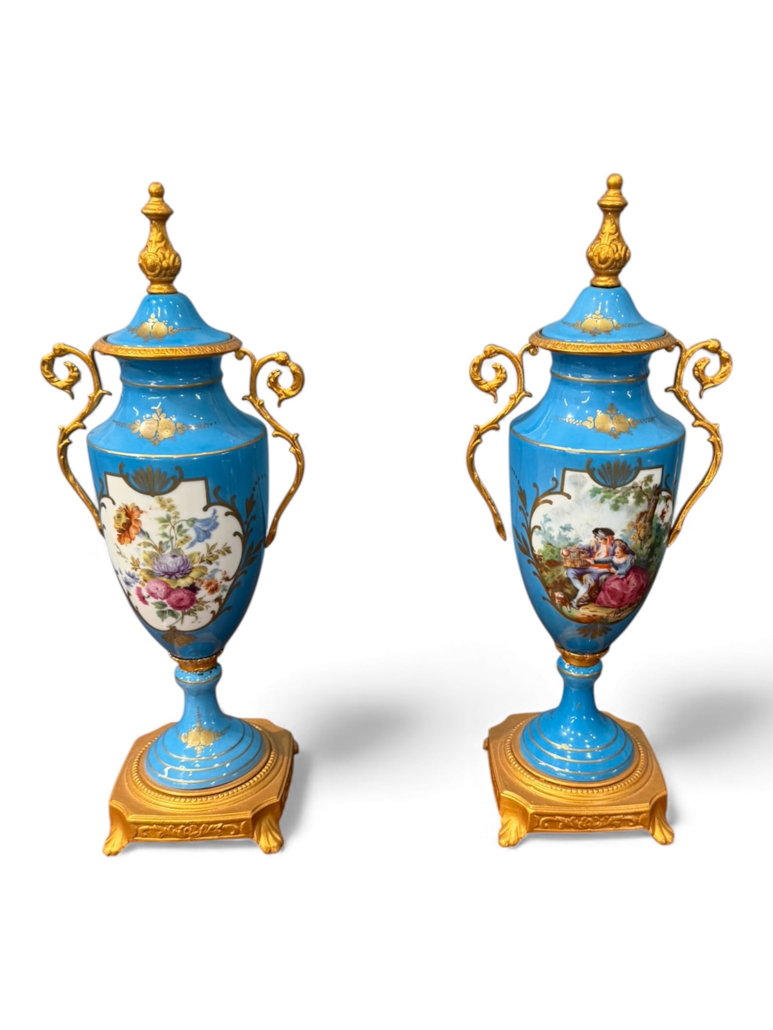 Pair of Gilt-Bronze Mounted Blue Porcelain Urns with Painted Panels, 20th century: A matched pair of decorative porcelain urns in vivid sky-blue glaze, each mounted with ornate gilt-bronze handles, finials, and square bases. The bodies are adorned with hand-painted reserves: one urn