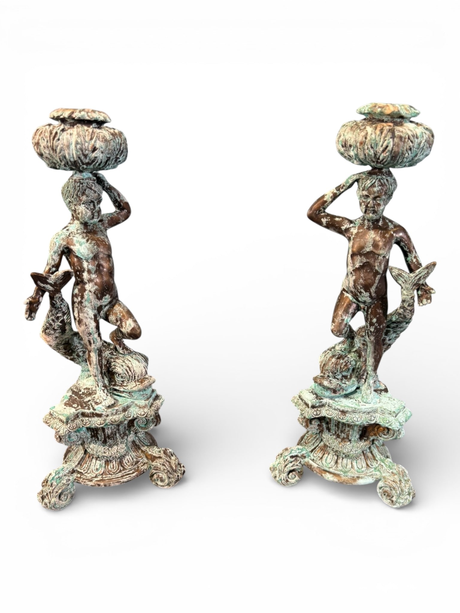 Pair of Figural Metal Candlesticks, Classical Style: A decorative pair of figural metal candlesticks modeled as standing male figures in the Classical taste, each supporting a foliate candle cup above the head. The figures are shown in contrapposto pose